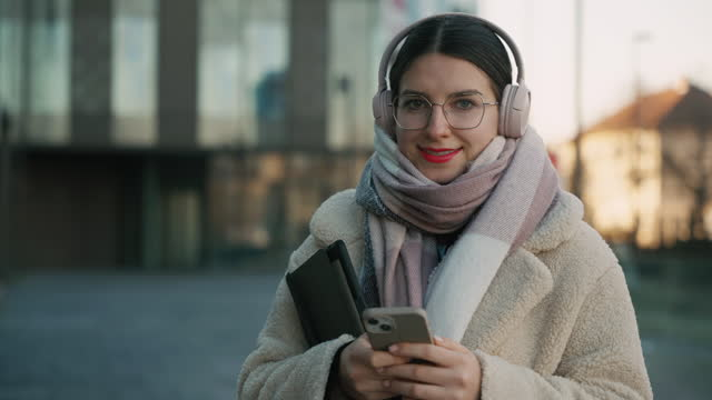 Женщина в наушниках и шарфе улыбается, держа телефон и планшет на улице.